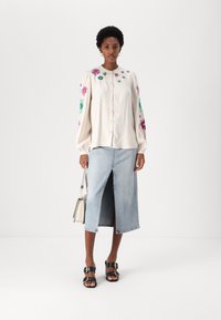Lys beige bluse med blomsterbroderi i grønt og rosa, kombinert med en lys blå denimskjørt med splitt foran. Svarte sandaler og en lys håndveske.