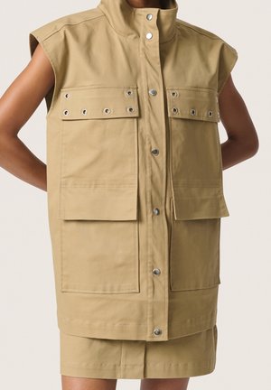 Ærmeløs beige jakke med høj krave og flere klaplommer, knaplukning foran, båret over matchende nederdel, arme bøjet bag ryggen.