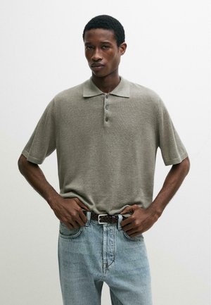 Uomo che indossa una polo grigia a maniche corte infilata in un paio di jeans azzurri con una cintura intrecciata marrone scuro, su uno sfondo semplice.