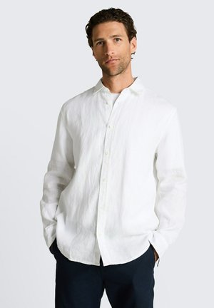 Mann mit lockigem dunklem Haar, der ein weißes Leinenhemd mit Knöpfen trägt und die Hände in den Taschen dunkler Hosen vor schlichtem Hintergrund hat.