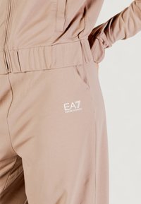 Chándal de color rosa claro con cintura elástica, que incluye bolsillos laterales y un pequeño logo blanco de EA7 en el muslo izquierdo. Textura suave.