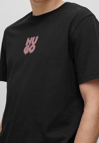 Sort bomulds T-shirt med et pink spray-stil trykt logo "HUGO" på brystet. Korte ærmer med rund halsudskæring.