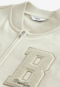 Lysbeige lynlåsjakke med ribbet krave, prydet med et tekstureret "B" logo i cremefarve på brystet og en sølvlynlås. Blødt stof i teksturen.