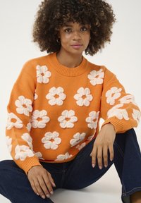 Narancssárga kötött pulóver fehér virágmintával, kerek nyakkivágással és bordázott mandzsettával. Sötétkék farmernadrággal párosítva, bemutatva egy laza stílust.