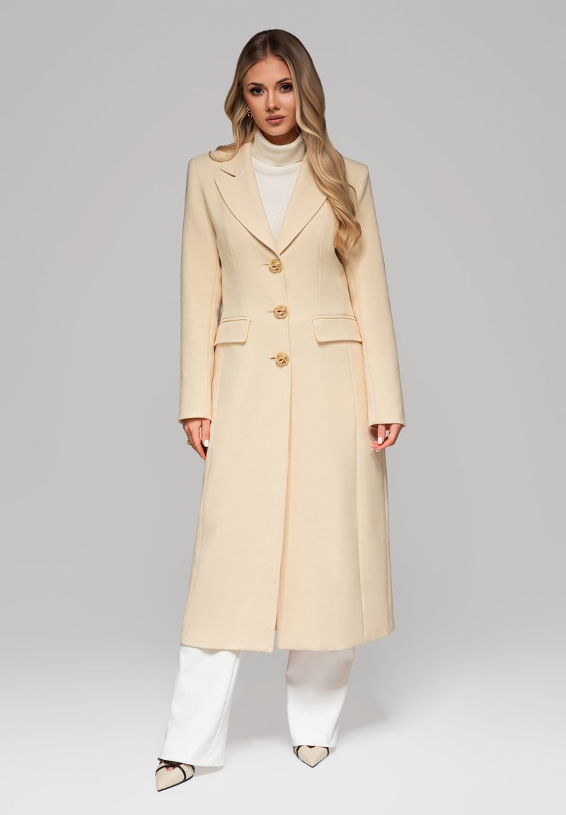 Lungo cappotto beige con un design su misura, caratterizzato da un colletto a revers, tre bottoni dorati e due tasche anteriori. Texture morbida e liscia.