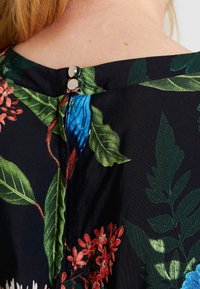 Gros plan sur une robe noire à motifs floraux avec des feuilles vertes, des fleurs rouges et bleues, maintenue au niveau du col arrière par deux boutons en argent.