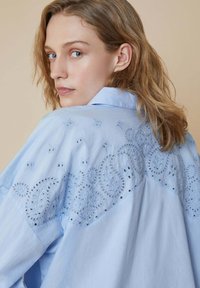 Maglietta azzurra con un design ricamato che presenta motivi elaborati e dettagli a occhiellini lungo la spalla e la schiena. Tessuto morbido.