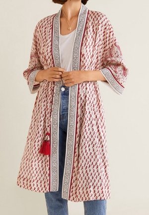 Kobieta nosząca sięgający kolan, wzorzysty kardigan w stylu kimono z czerwonymi i szarymi obramowaniami, na białej koszuli i niebieskich dżinsach.