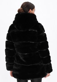 Cappotto in pelliccia sintetica nera con cappuccio, caratterizzato da una consistenza spessa e soffice e un design trapuntato. La silhouette è morbida e di lunghezza sotto la vita.