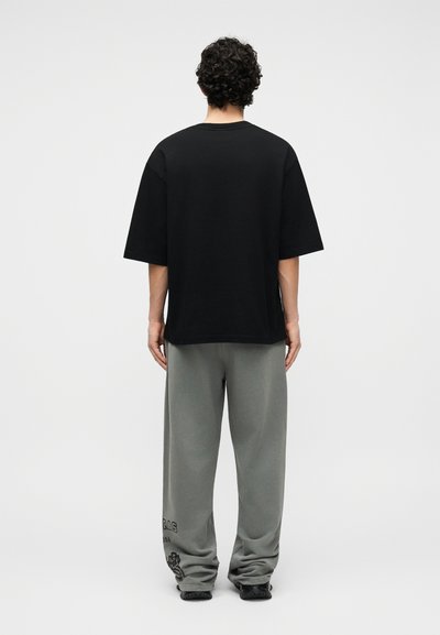 Άνδρας με σγουρά μαλλιά στέκεται γυρισμένος προς τα πίσω, φοράει μια φαρδιά μαύρη μπλούζα και γκρι φόρμα με μαύρα σχέδια κοντά στον αριστερό αστράγαλο.