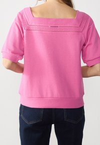 Žena v růžovém tričku s krátkým rukávem a čtvercovým výstřihem, zdobeném kraječkami, ve spojení s tmavě modrými denimovými džíny.