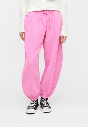 Άτομο που φορά χαλαρό ροζ παντελόνι με κορδόνι και ελαστικά μανσέτες, καθώς και μαύρα ψηλά αθλητικά παπούτσια, στέκεται μπροστά σε λευκό φόντο.