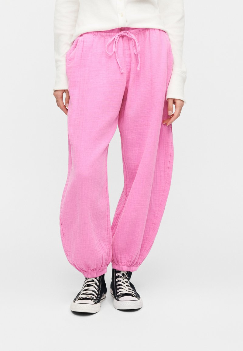 Άτομο που φορά χαλαρό ροζ παντελόνι με κορδόνι και ελαστικά μανσέτες, καθώς και μαύρα ψηλά αθλητικά παπούτσια, στέκεται μπροστά σε λευκό φόντο.