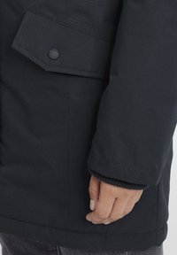 Chaqueta azul marino oscura con un acabado mate, que presenta una solapa abotonada en la manga y puños acanalados. Detalle de costura visible a lo largo de las costuras.
