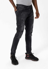 Homme portant un jean noir slim déchiré et des baskets noires, debout avec une main dans la poche devant un fond blanc.