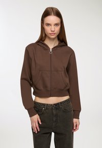 Giovane donna che indossa un cappotto corto di lana marrone con zip e jeans neri a vita alta, in piedi davanti a uno sfondo bianco semplice.