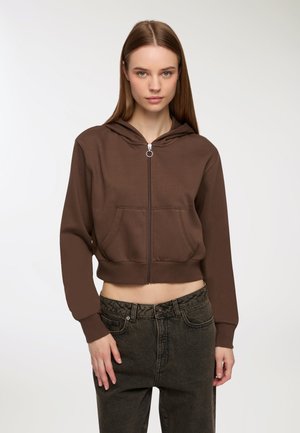 Giovane donna che indossa un cappotto corto di lana marrone con zip e jeans neri a vita alta, in piedi davanti a uno sfondo bianco semplice.