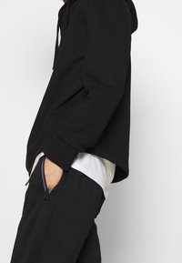 Schwarze Hoodie mit lockerer Passform, ausgestattet mit einer Kängurutasche und einer verstellbaren Kapuze mit Kordelzug; der Stoff wirkt weich und glatt.