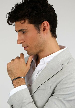 Grauanzugjacke über einem weißen Hemd, mit einem schwarz-silbernen Armband am Handgelenk. Die Haare sind lockig, im Profil zu sehen.