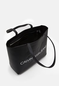 Svart tygväska av slät material med två långa remmar. Har en zip-ficka och "CALVIN KLEIN JEANS"-logotyp på framsidan.