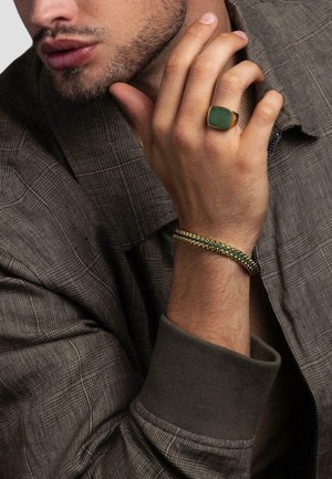 Gold ring with green stone, layered chain bracelet featuring green accents, and a textured brown patterned shirt with a ribbed collar.