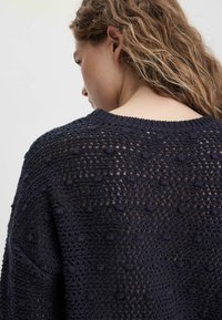 Marineblauer gestrickter Pullover mit offenem Webmuster, verziert mit Knötchenakzenten, runder Halsauschnitt und lockerer Passform. Die Rückansicht zeigt die Nahtdetails.