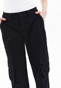 Zwarte cargobroek met knoopsluiting, zij- en voorzakken, en een relaxte pasvorm van textuurstof met contrasterende zijnaad.