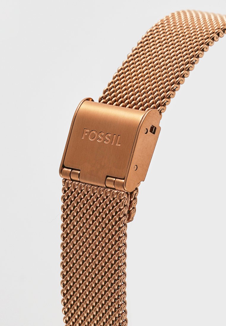 Różowe złoto, metalowa bransoleta w siatkę z zapięciem składanym, wygrawerowanym logo marki "FOSSIL" na jasnym tle.