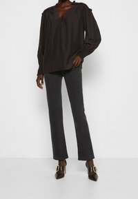 Blouse noire à manches longues avec un col en V et un détail noué, présentant des poignets élastiques texturés ; portée avec un pantalon droit foncé et des chaussures à talons.