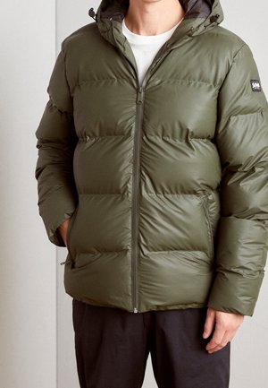 Mężczyzna ubrany w oliwkowo-zieloną pikowaną kurtkę z przednim zamkiem błyskawicznym i bocznymi kieszeniami, zestawioną z czarnymi spodniami i białą warstwą pod spodem.