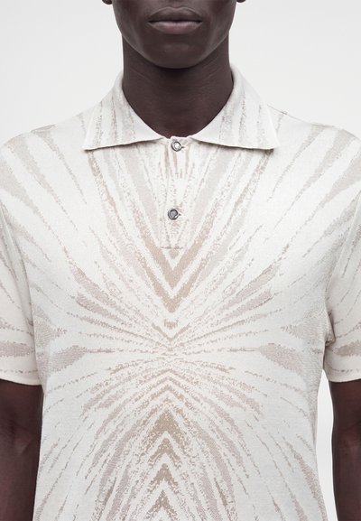 Personne à la peau foncée portant un polo crème à manches courtes avec un motif symétrique en forme d'éclat de soleil brun clair et deux boutons au col.