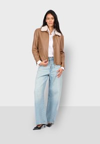 Bruine leren bomberjack met een witte schapenvachten kraag, rits sluiting, gecombineerd met lichtblauwe wijdvallende jeans en zwarte hakken.