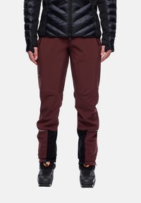 Person in burgunderfarbenen und schwarzen Outdoor-Hosen, schwarzen Skistiefeln und einer schwarzen gesteppte Jacke, sichtbar von der Taille bis zu den Füßen.