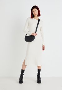 Vestido branco de manga longa e canelado com fenda lateral. Usado com uma mala preta de tiracolo e botas pretas de salto grosso, em um fundo liso.