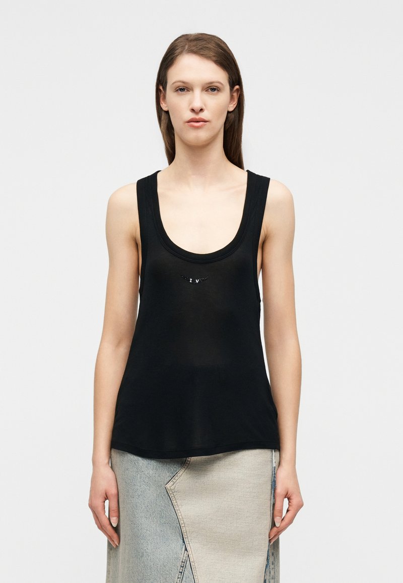 Νεαρή γυναίκα με ίσια καστανά μαλλιά, φορώντας μ αύρο αμάνικο τοπ και δίχρωμη τζιν φούστα, στέκεται μπροστά σε λευκό φόντο.