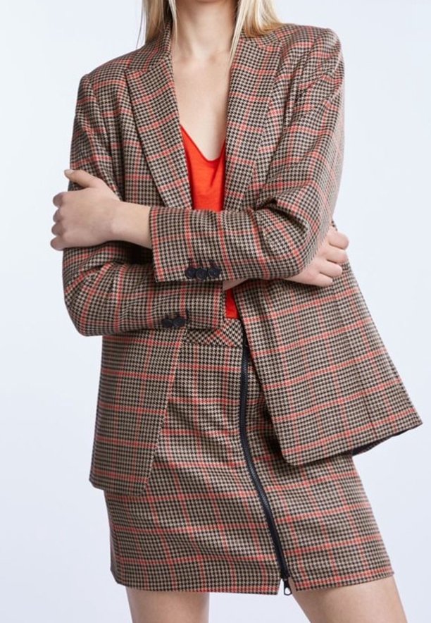 Gecontroleerde blazer in bruin en rood met gewatteerde schouders, zwarte knopen, gedragen over een rode top; gecombineerd met een bijpassende, nauwsluitende ritsrok.