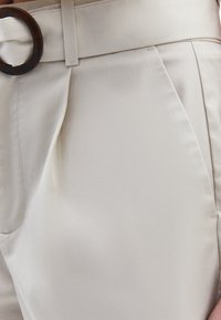 Pantalons beige clair avec une texture lisse. Comprend une large ceinture et une boucle de ceinture ronde en marron foncé. Coupe ajustée avec des plis.