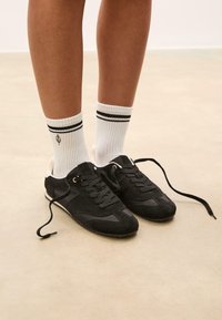 Sorte atletiske sko med ruskind og glatte teksturer, der har sorte snørebånd og en hvid sål, parret med hvide sokker prydet med sorte striber.