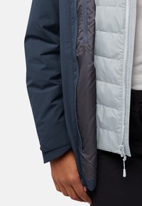 Veste zippée avec une couche extérieure bleu marine, une doublure intérieure en matelassage gris clair et une texture lisse et mate. Comprend une poche visible.