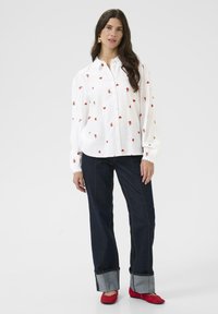 Witte button-up shirt met rode hart- en bloemenborduursels, gestyled met donkere denim broek met een omgeslagen zoom en rode schoenen.