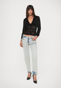 Woman standing with left hand in light blue jeans pocket, wearing black floral lace buttoned shirt and black high-heeled shoes.