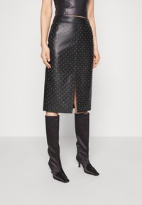 Femme portant une jupe crayon en cuir noir avec des clous métalliques, une fente à l'avant, et des bottes à talons hautes jusqu'aux genoux noires, debout contre un fond uni.