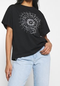 Camiseta de algodón negra con un gráfico blanco de un sol y un diseño de cuadrícula, corte relajado con mangas cortas, combinada con jeans de color azul claro.