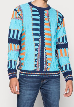 Homme portant un pull en maille coloré avec des rayures verticales et horizontales bleues, orange et marine, assorti à un jean bleu.
