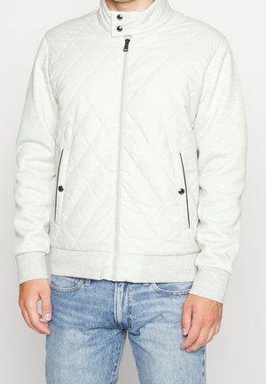 Mann trägt eine weiße gesteppte Jacke mit Reißverschluss, Druckknopfkragen und Seitentaschen, kombiniert mit hellblauen Jeans.