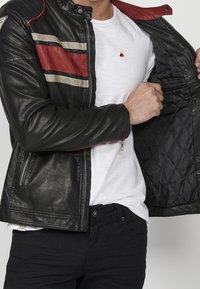 Uomo con giacca di pelle nera con strisce rosse e beige, che apre la giacca per mostrare la fodera interna imbottita, t-shirt bianca e pantaloni neri.
