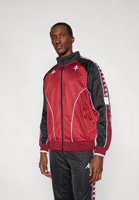 Bordowa i czarna kurtka zapinana na zamek z białymi akcentami, ściągaczami na mankietach i detalami z logo. Posiada stójkę i boczne paski.