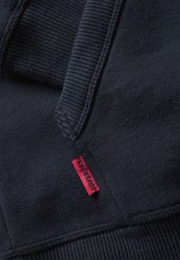 Granatowy sweter z ściągaczami przy rękawach i dole, z małą czerwoną metką z napisem "Superdry". Miękka tekstura i minimalistyczny design.