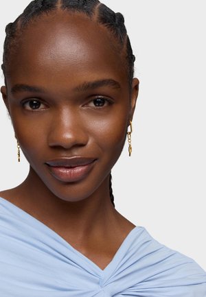 Helles blaues Oberteil mit rüschiger Textur, getragen von einer Person mit geflochtenem Haar und goldenen Kreolen, die ein natürliches Make-up trägt.