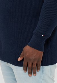 Ναυτικό πλεκτό πουλόβερ με ριγωτή υφή, διαθέτοντας ένα μικρό κόκκινο, λευκό και μπλε λογότυπο στο μανίκι. Φοριέται πάνω από ανοιχτό μπλε τζιν.